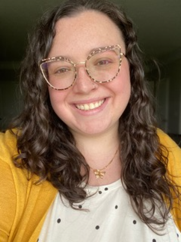 Picture of Kennedy Sepsi. Woman with printed glasses smiling. Yellow cardigan with white and black poka dot shirt under. 