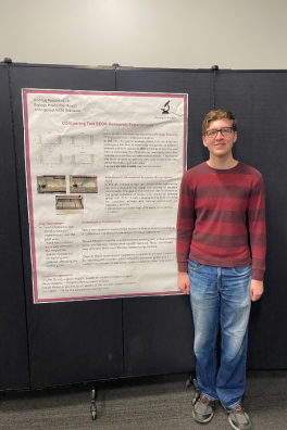 Male student wearing red striped shirt and jeans smiling and standing in front of research poster.