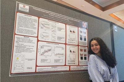 Student with long dark hair in a striped shirt smiling and standing in front of research poster.