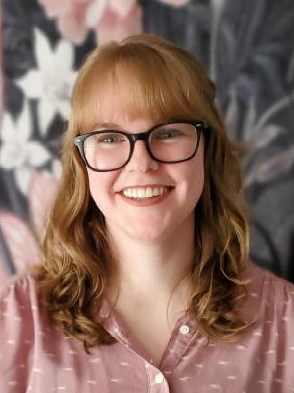 Rebecca is wearing glasses and a pink shirt standing in front of a floral background.