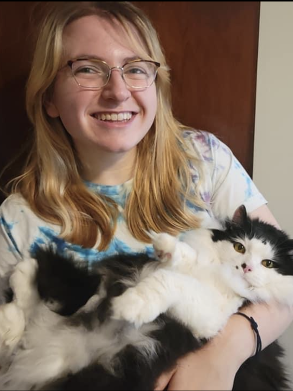 Picture of Maddy McCain (they are a blonde haired person, with glasses, holding a black and white cat while smiling)